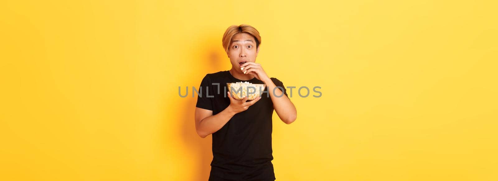 Excited and amazed asian guy watching interesting movie and eating popcorn from bowl, standing yellow background.