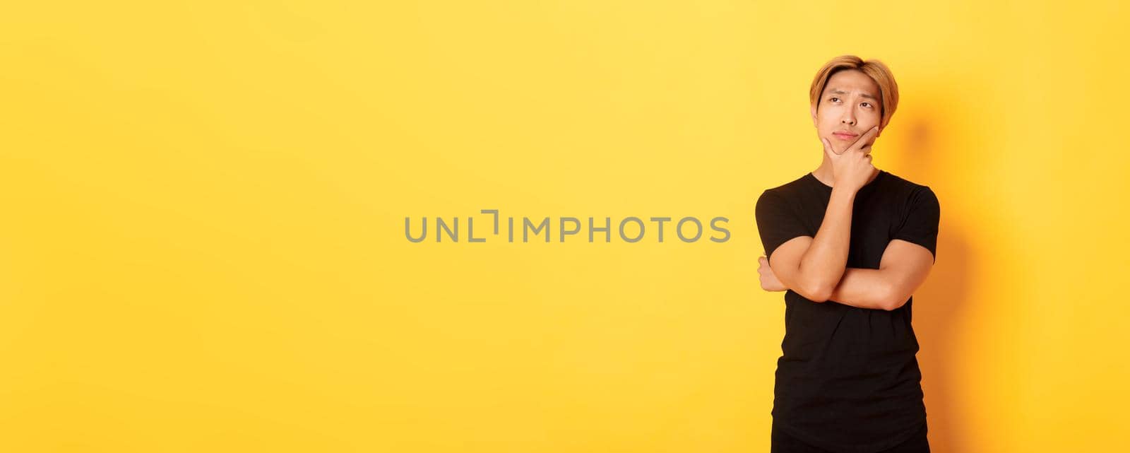Portrait of thoughtful attractive guy in black t-shirt looking upper left corner. Young man pondering idea, standing yellow background by Benzoix