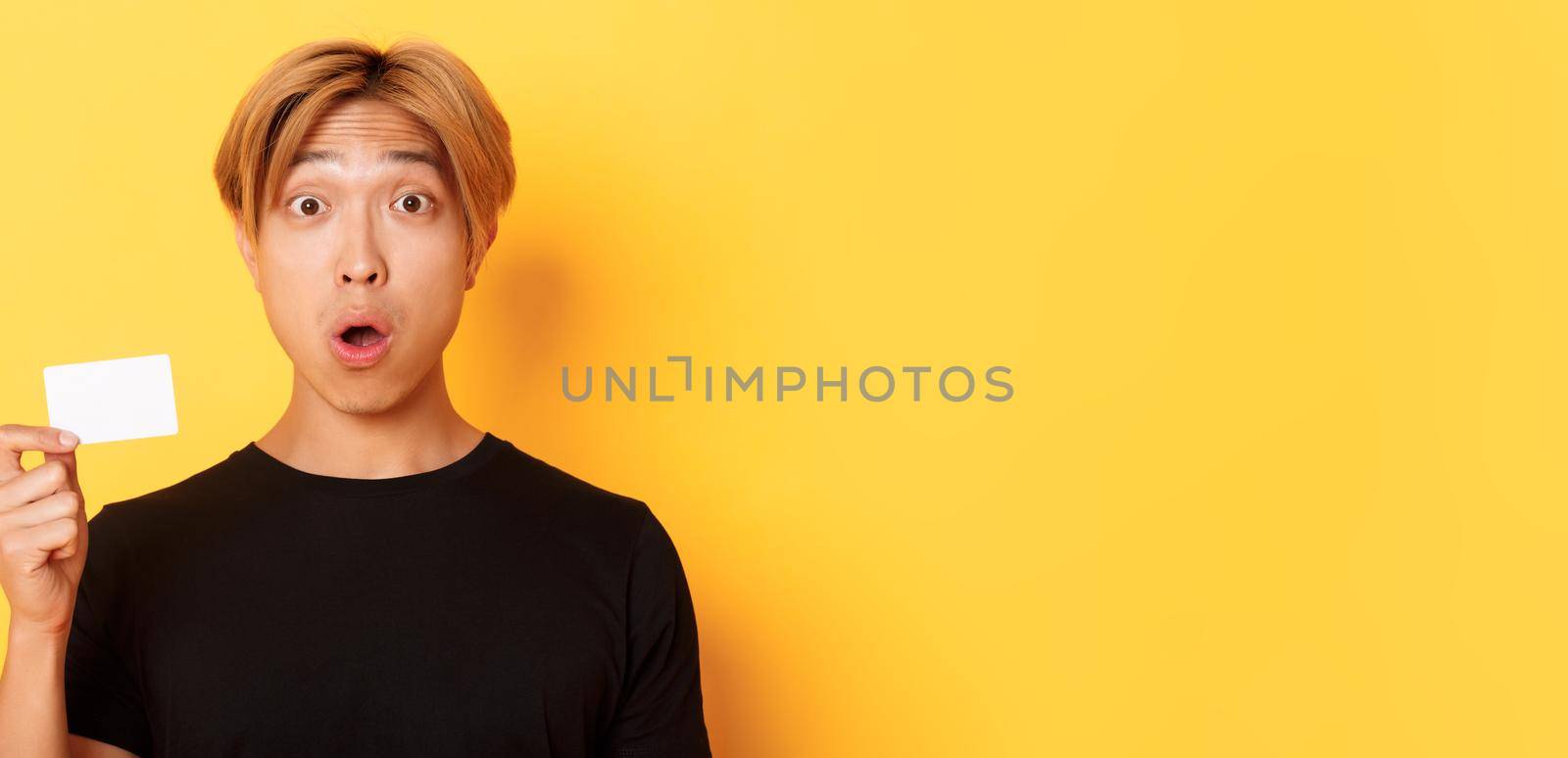 Close-up of surprised gasping asian guy with fair hair, looking amazed and startled as showing credit card, standing yellow background by Benzoix