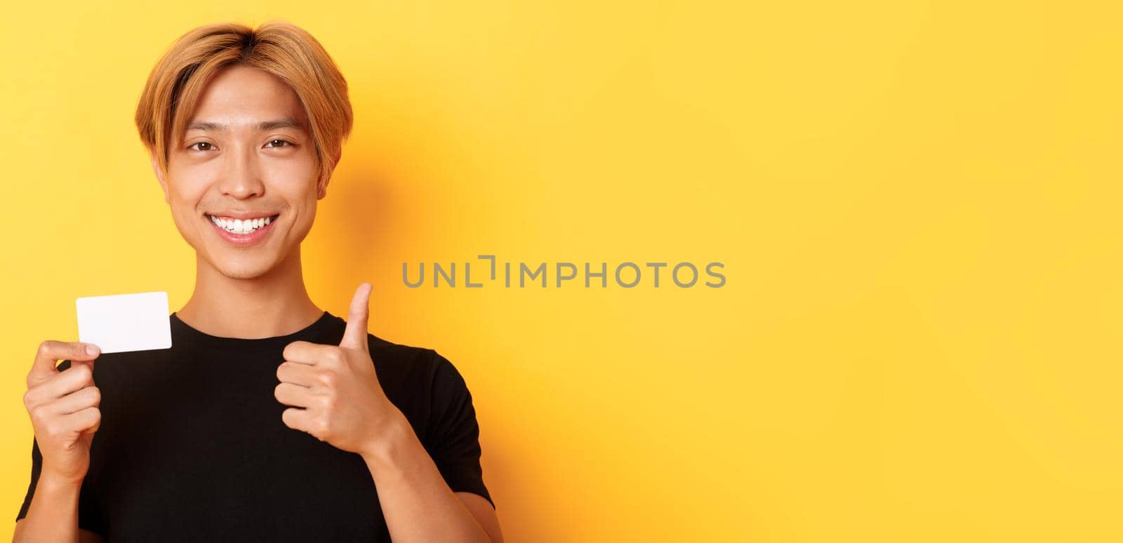 Close-up of satisfied asian guy recommend bank, showing credit card and standing over yellow background by Benzoix