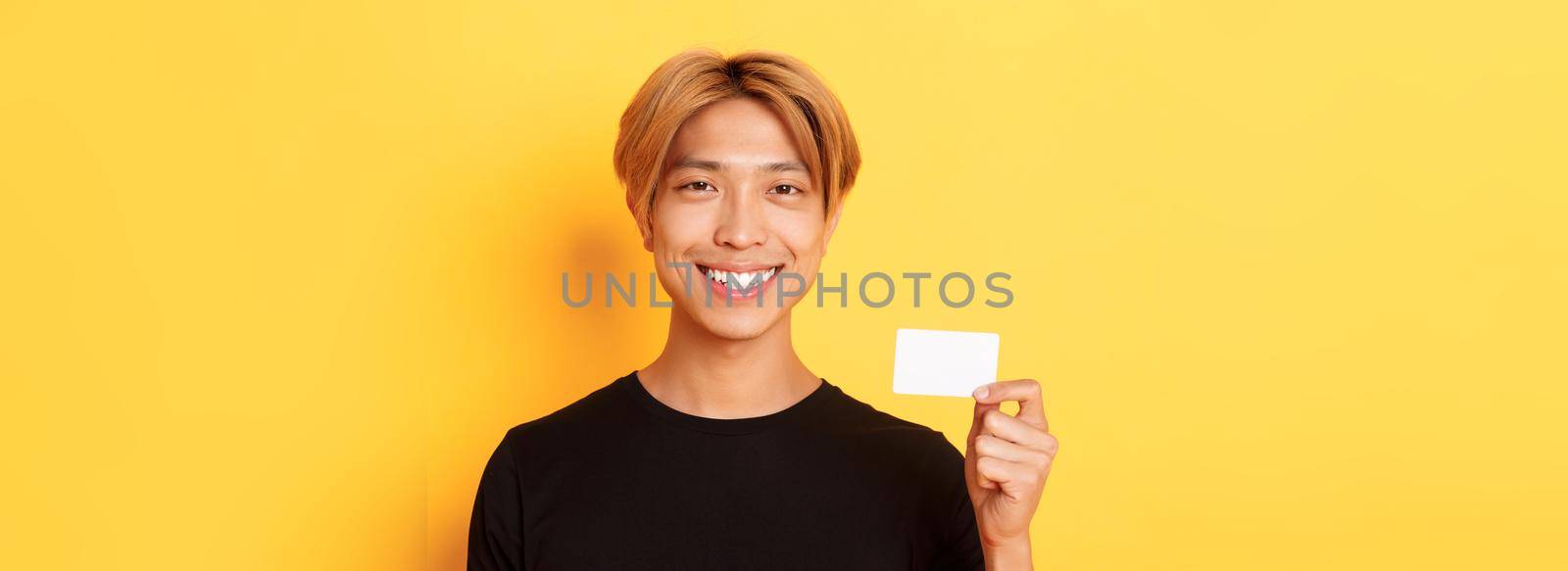 Close-up of handsome pleased asian guy with fair hair, smiling happy and showing credit card over yellow background.