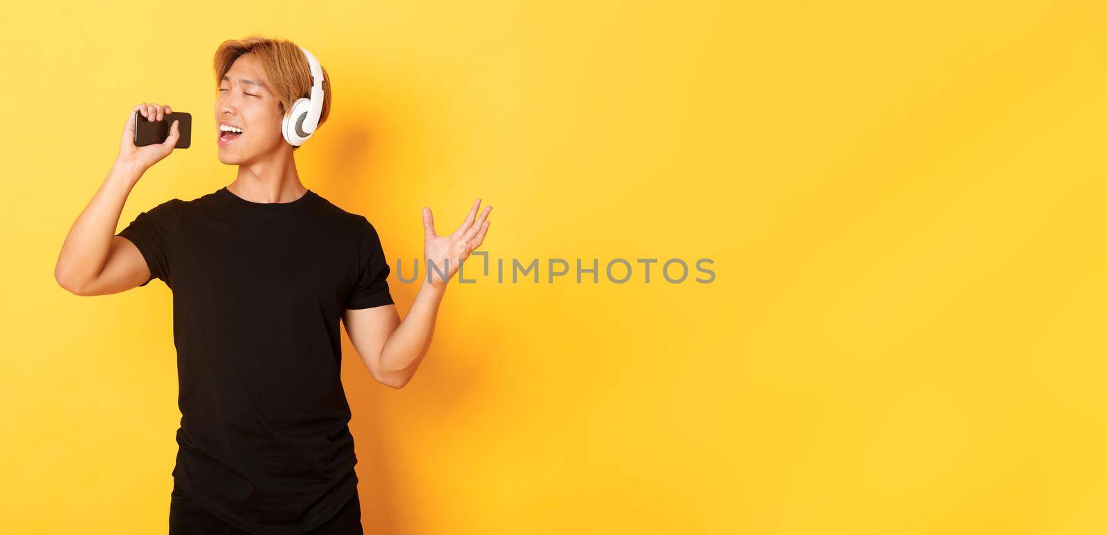 Joyful attractive korean guy in headphones, playing karaoke app, singing into mobile phone microphone, standing yellow background by Benzoix
