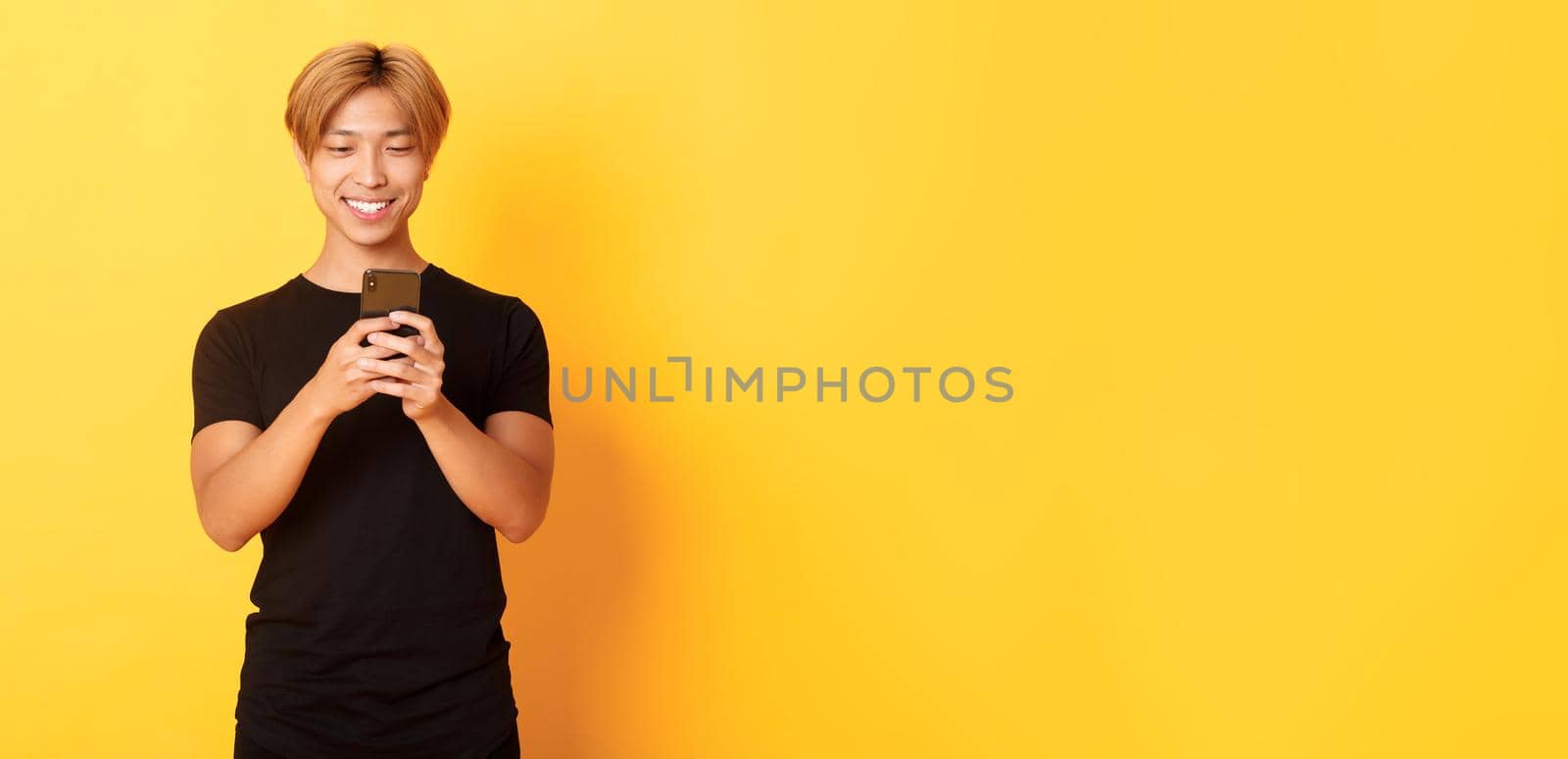 Portrait of handsome stylish asian guy with blond hair, using mobile phone and smiling, yellow background by Benzoix