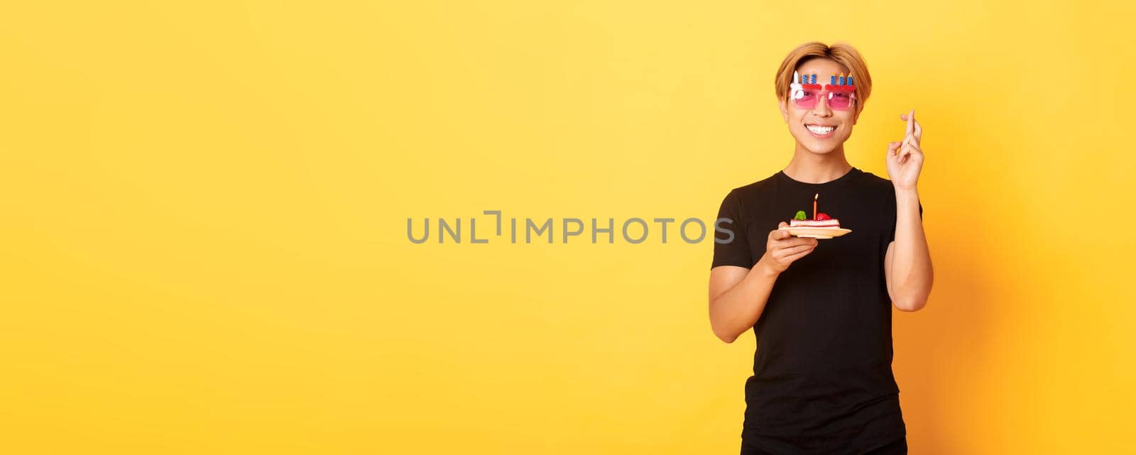 Portrait of hopeful attractive asian birthday guy, celebrating in funny party glasses, holding cake and fingers crossed while making b-day wish, yellow background by Benzoix