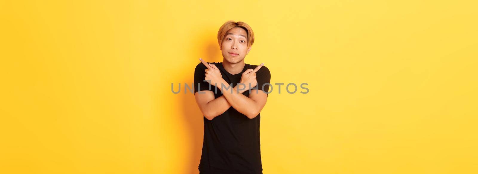 Indecisive handsome guy shrugging puzzled and pointing fingers sideways, standing yellow background.