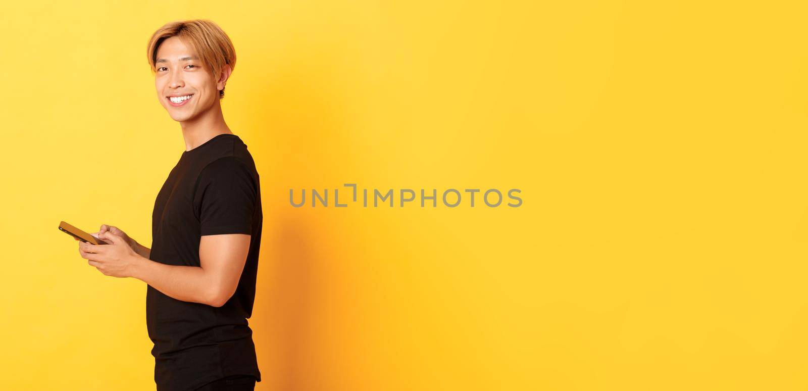 Portrait of handsome stylish asian guy in black outfit, using mobile phone and turning head at camera with satisfied smile, yellow background.