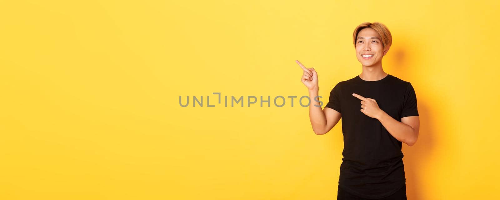 Portrait of handsome korean blond guy, pointing fingers upper left corner and looking satisfied with pleased smile, yellow background by Benzoix