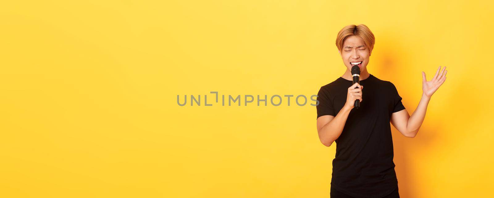 Handsome asian male singer, korean guy singing song at karaoke in microphone with passion, standing over yellow background.