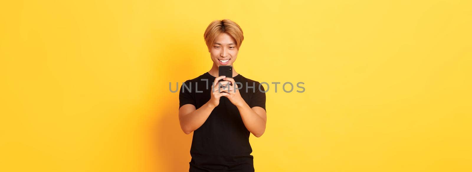 Portrait of handsome stylish asian guy with blond hair, using mobile phone and smiling, messaging in smartphone app, yellow background.