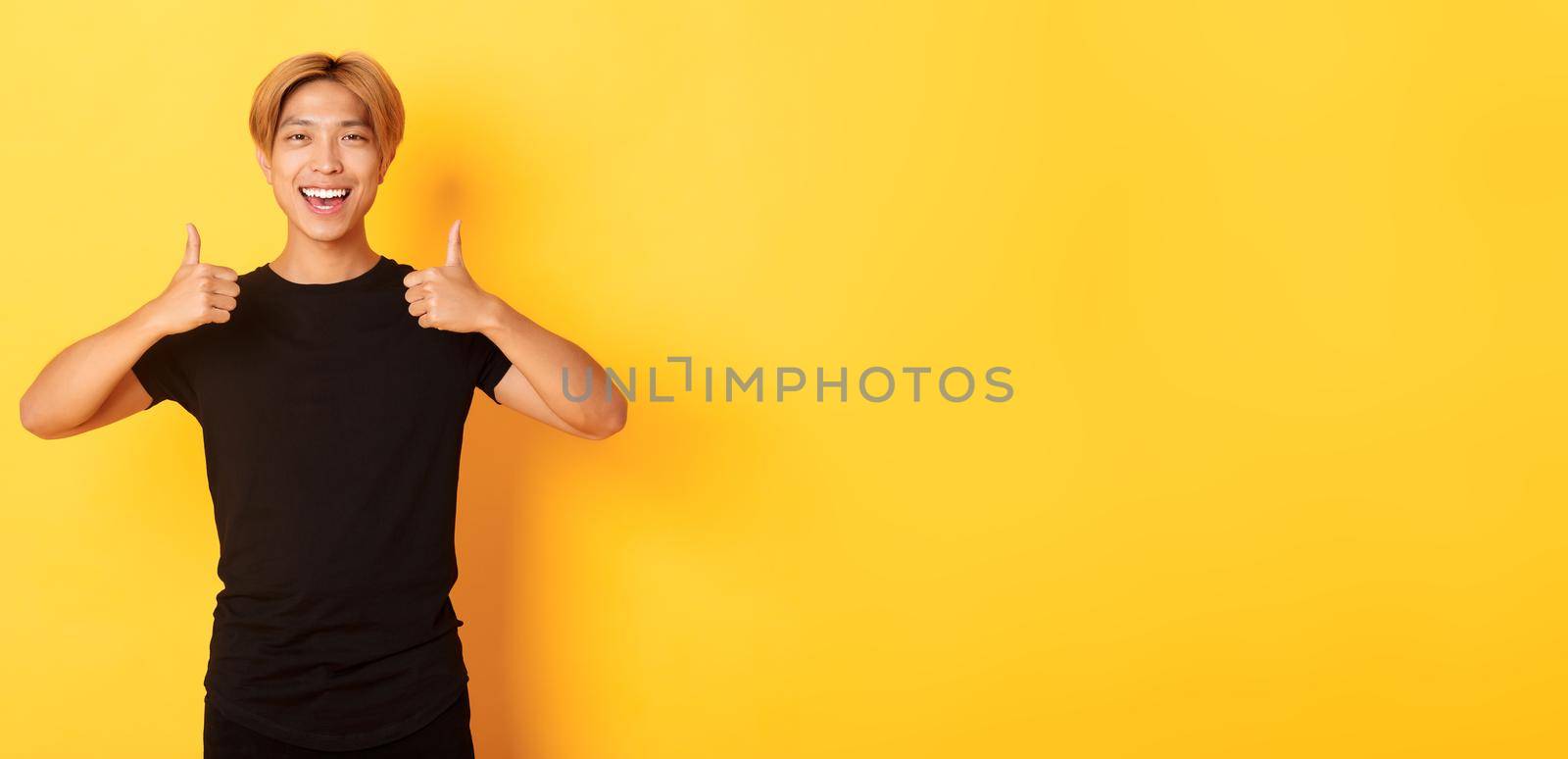 Portrait of satisfied handsome asian male student showing thumbs-up in approval, standing yellow background.