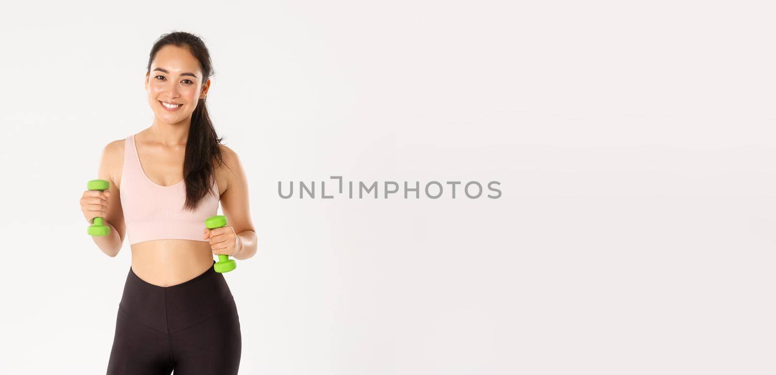 Fitness, healthy lifestyle and wellbeing concept. Portrait of happy slim and strong asian female athlete, sportswoman in activewear holding dumbbells for workout, exercise in gym, white background.