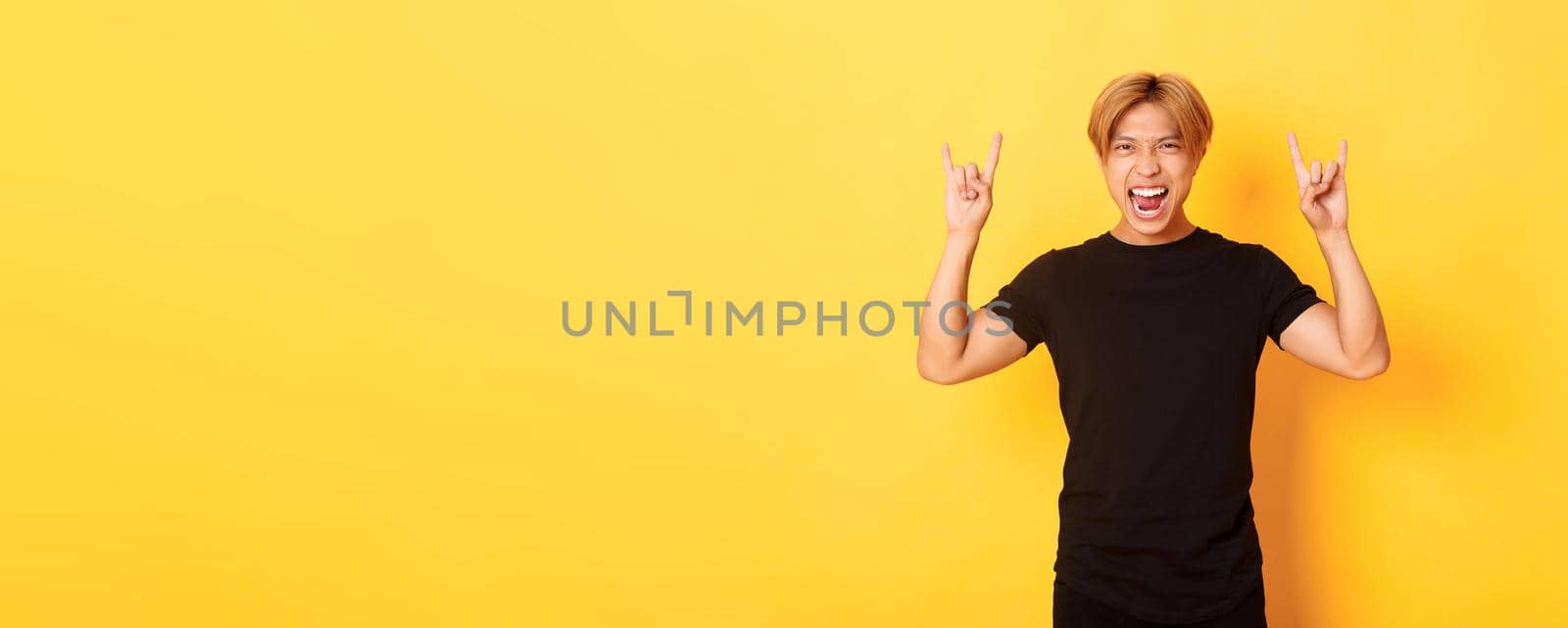 Portrait of happy handsome asian man having fun and partying, showing rock-n-roll gesture, standing yellow background by Benzoix