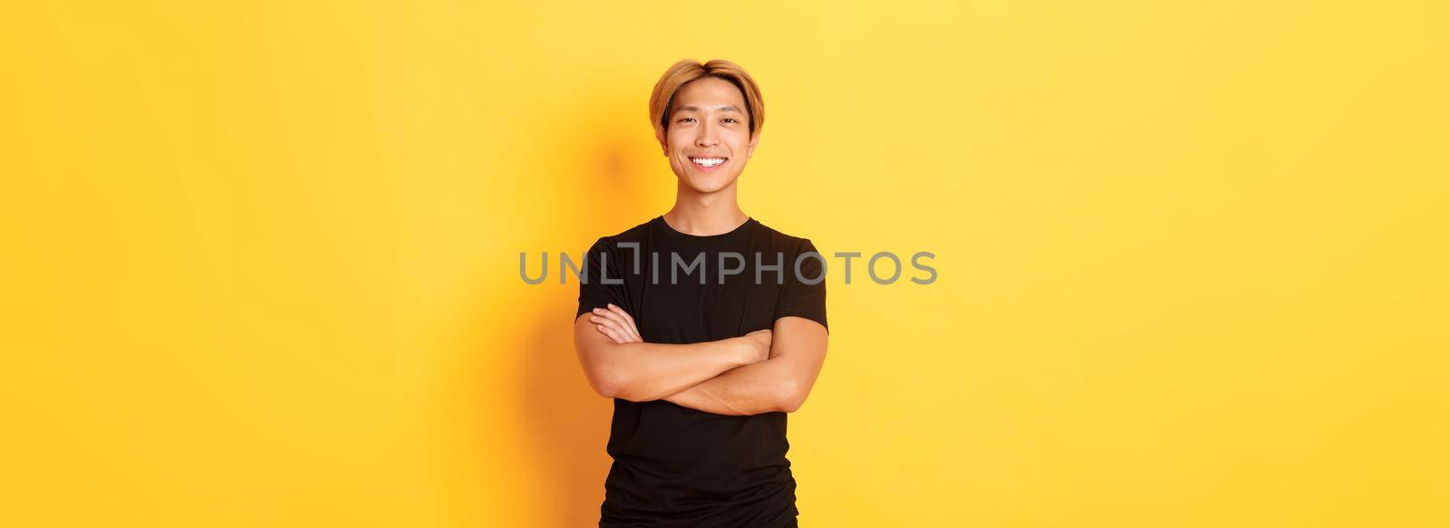 Portrait of confident handsome asian man smiling satisfied, standing over yellow background in black clothes.