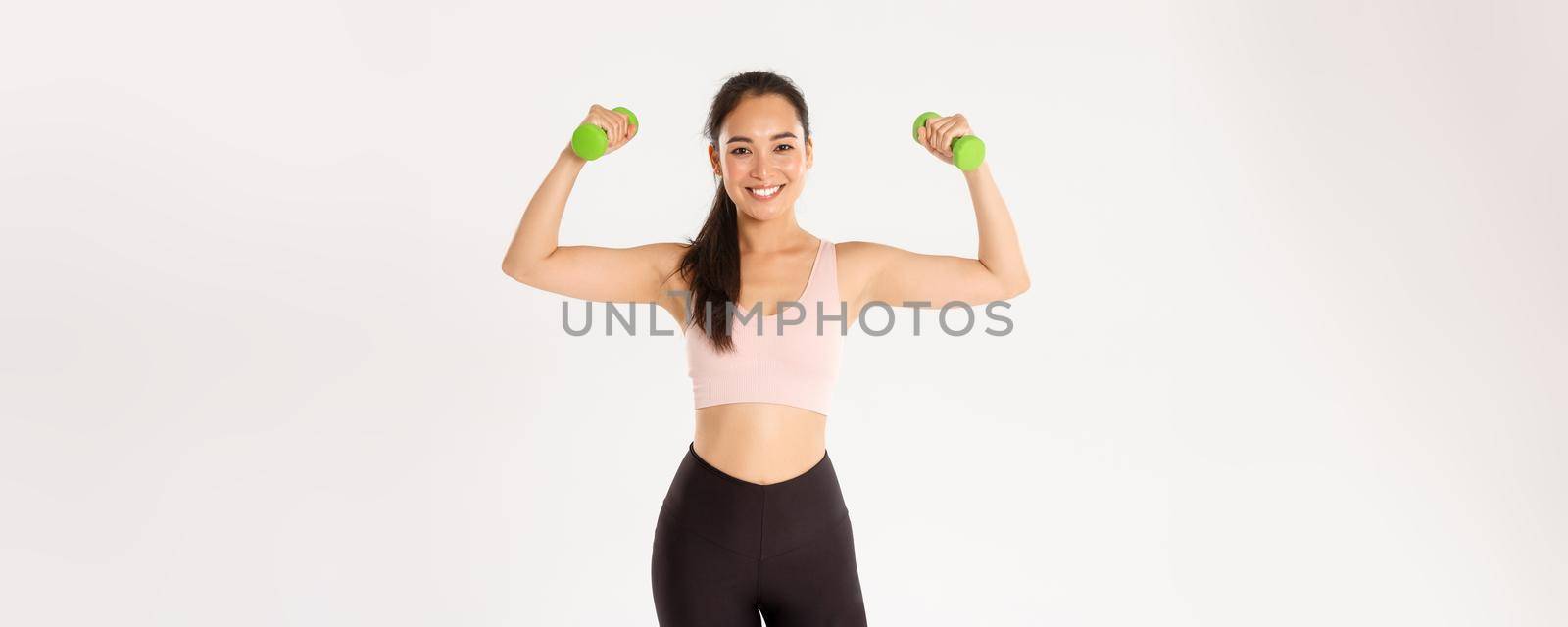 Fitness, healthy lifestyle and wellbeing concept. Portrait of strong and happy female athlete, asian girl workout at home during coronavirus, lifting dumbbells to gain muscles, white background.