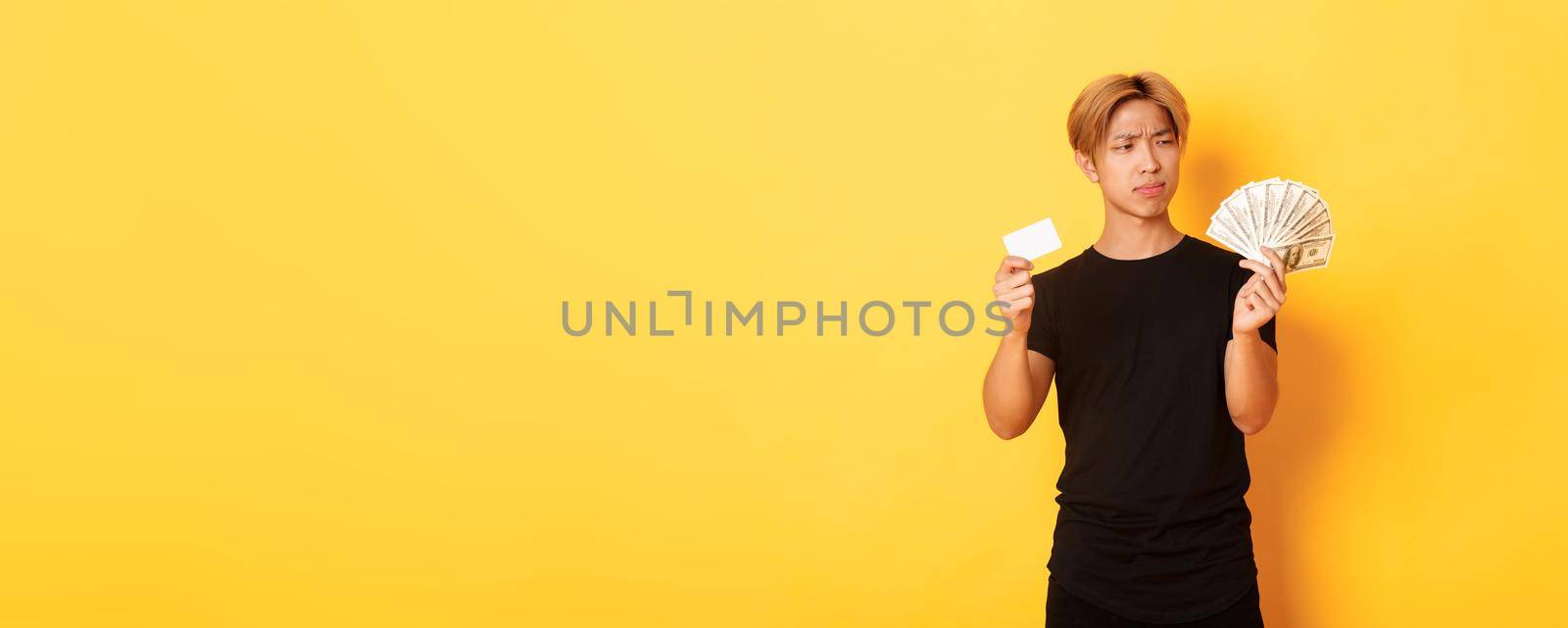 Indecisive and confused young asian guy looking puzzled at money while holding cash and credit card, yellow background.