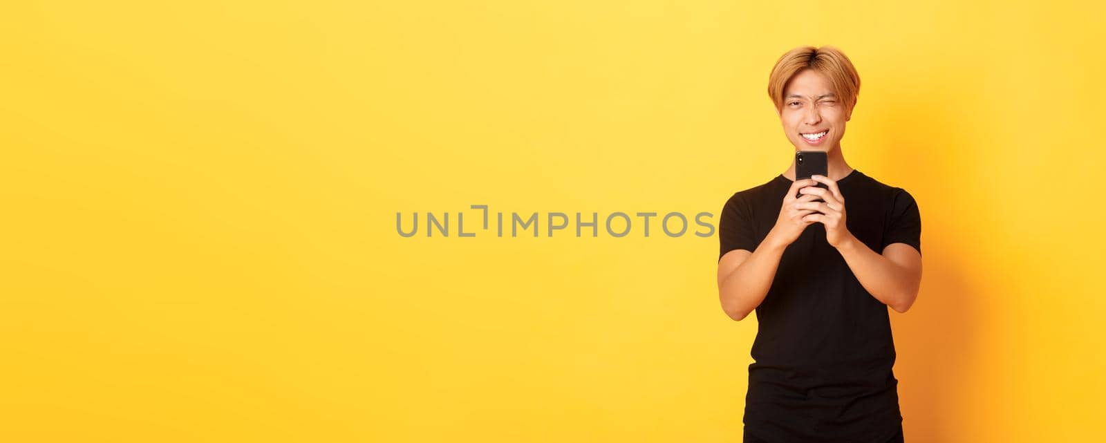 Stylish handsome asian guy taking picture on smartphone and smiling, photographing with mobile phone, standing yellow background by Benzoix