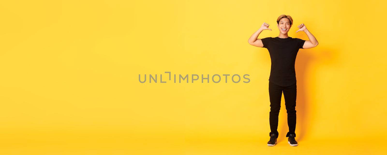 Confident attractive blond asian guy, smiling sassy and pointing at himself, standing over yellow background.