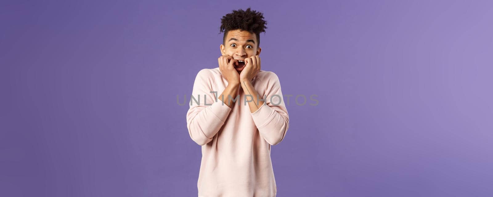 Portrait of young scared guy watching really scary movie, horror alone, gasping screaming and hold hands pressed to face, look terrified, trembling from fear, standing purple background.