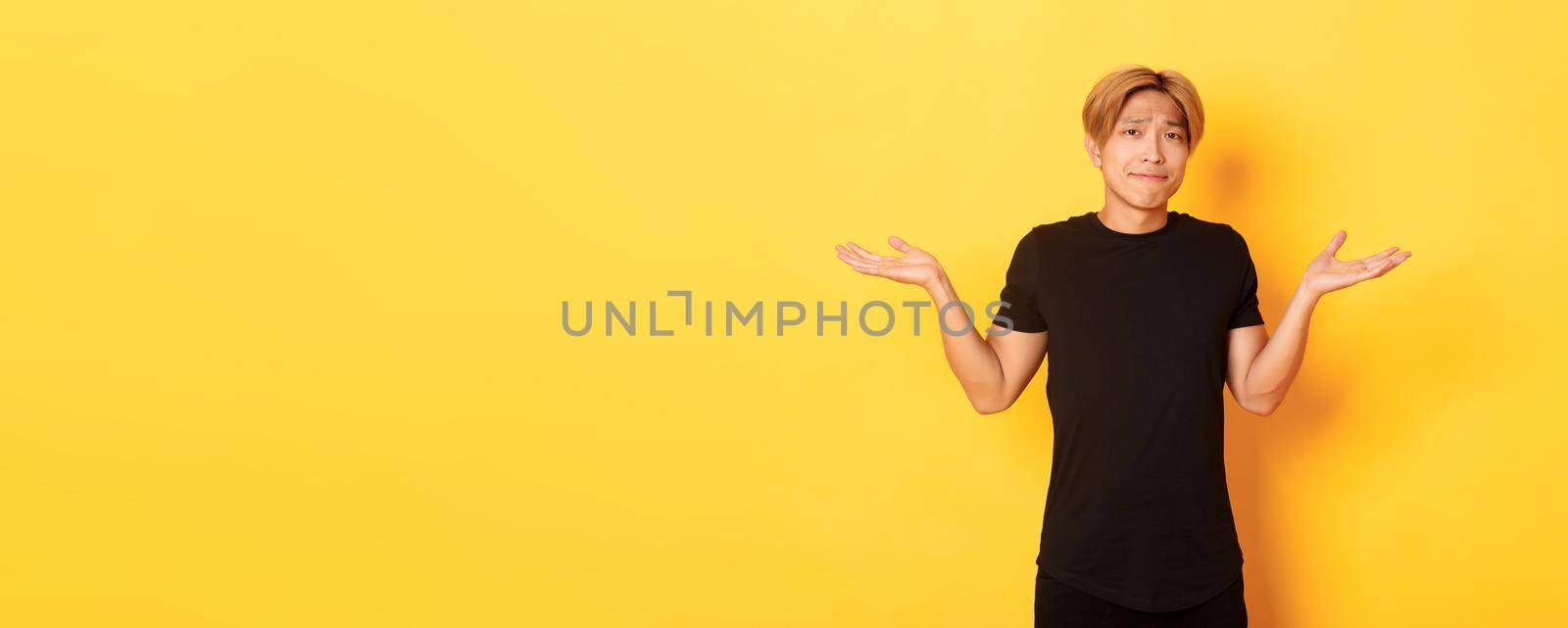 Portrait of confused and indecisive attractive korean guy, shrugging and pouting upset, standing yellow background.