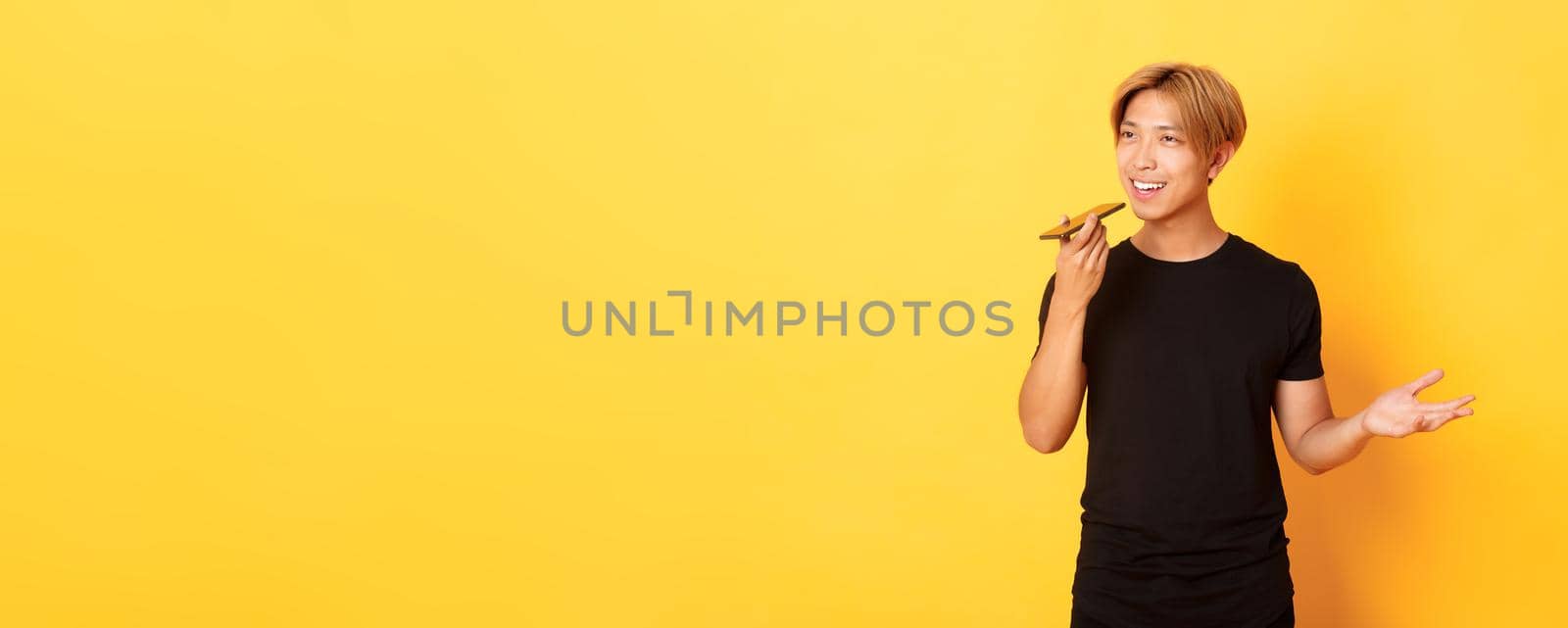 Portrait of stylish handsome korean guy with blond hair record voice message on mobile phone, holding smartphone near mouth and speaking, standing yellow background.