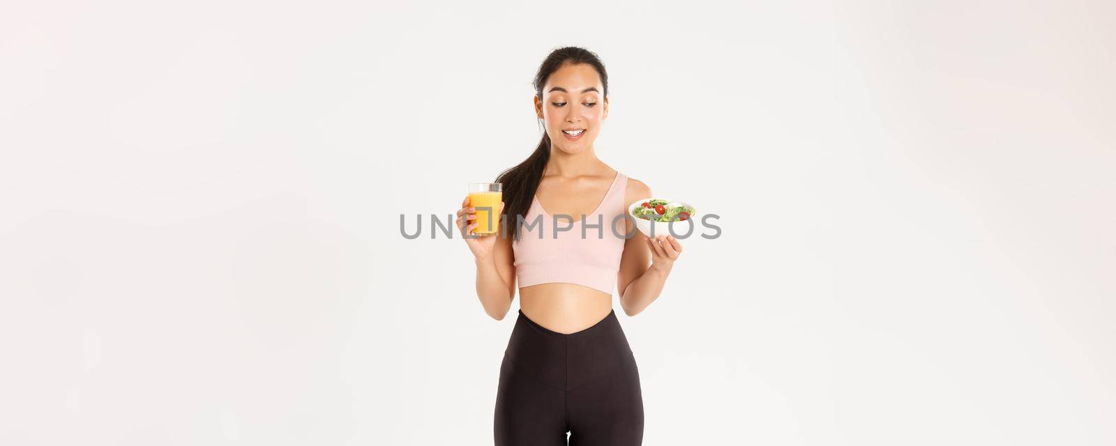 Sport, wellbeing and active lifestyle concept. Smiling healthy and slim brunette asian girl like fitness, going to gym and being on diet, holding salad with orange juice, standing white background by Benzoix