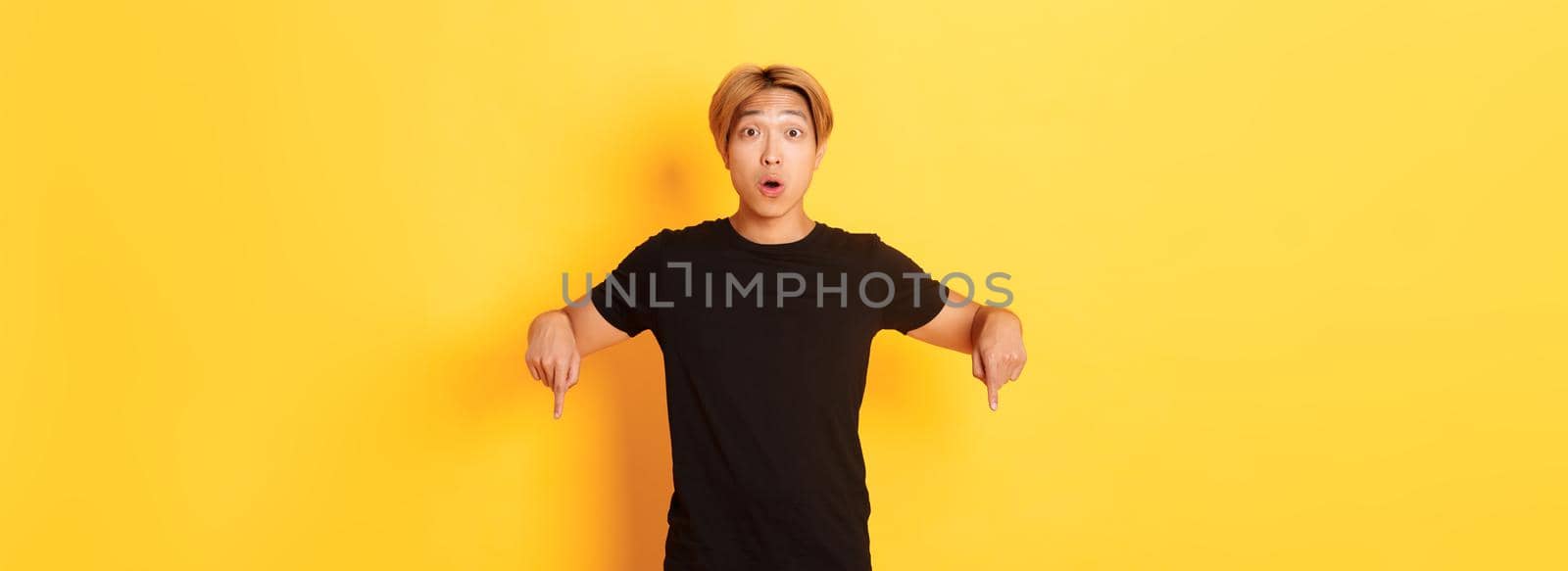 Amused happy asian guy, pointing fingers down and gasping impressed, showing awesome advertisement, yellow background.