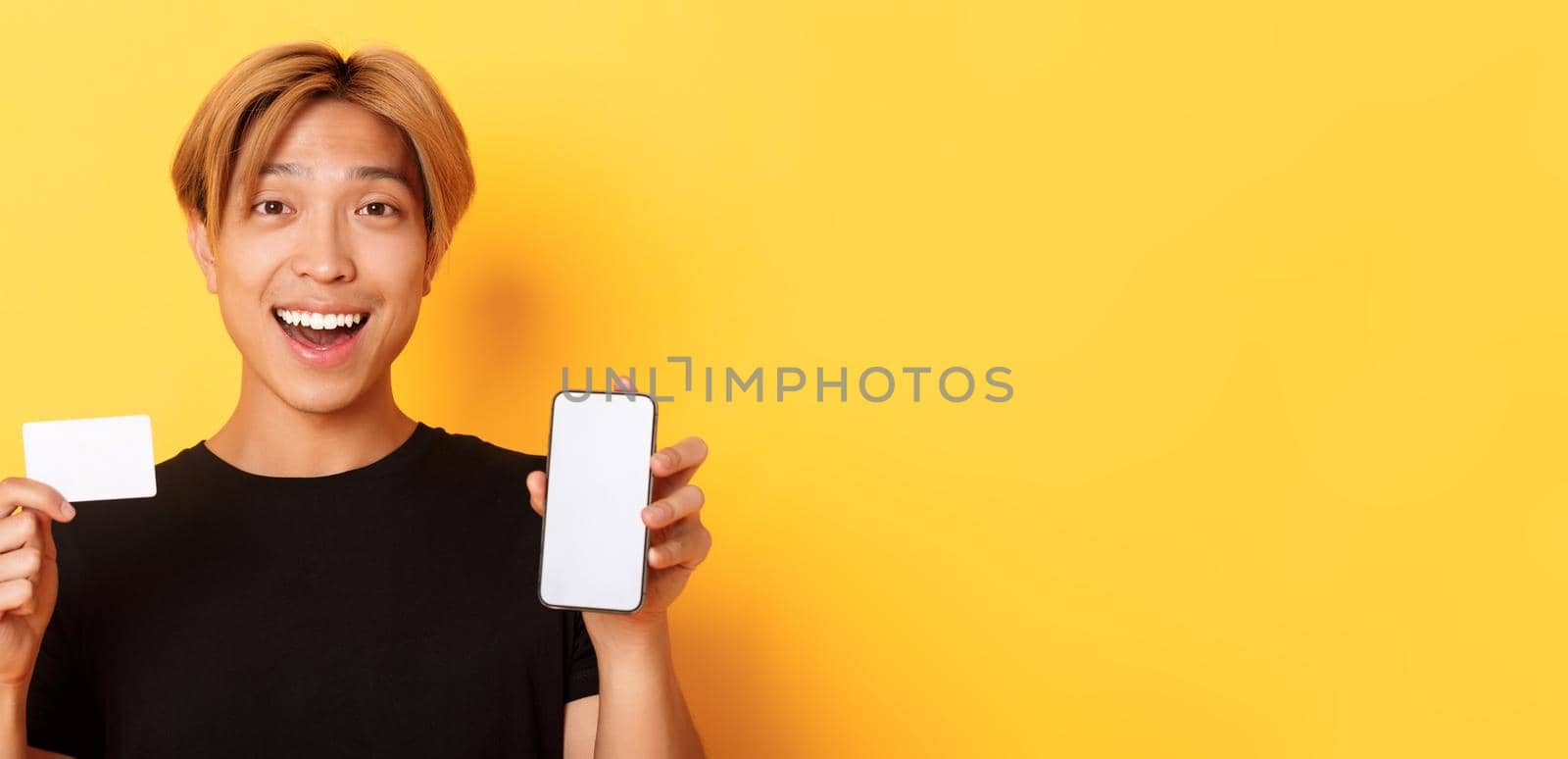 Amazed happy asian guy showing credit card and smartphone screen, smiling fascinated, standing yellow background.