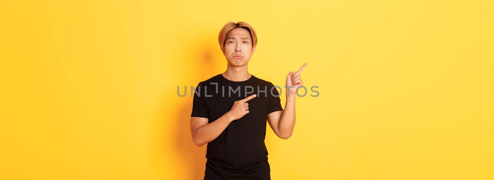 Portrait of misearble and gloomy asian guy in black t-shirt, pointing fingers upper left corner disappointed, yellow background by Benzoix