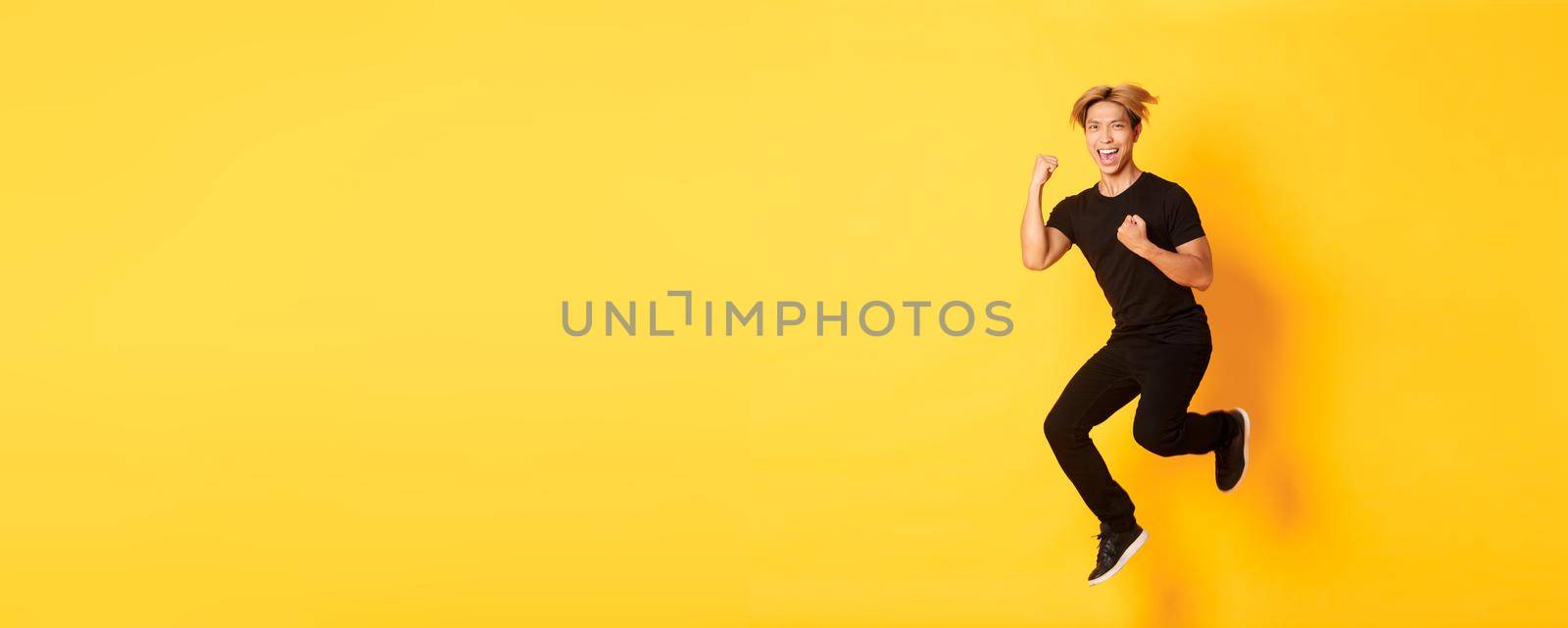 Full-length of happy attractive asian guy in black clothes jumping and celebrating victory, achieve goal, standing yellow background, triumphing by Benzoix