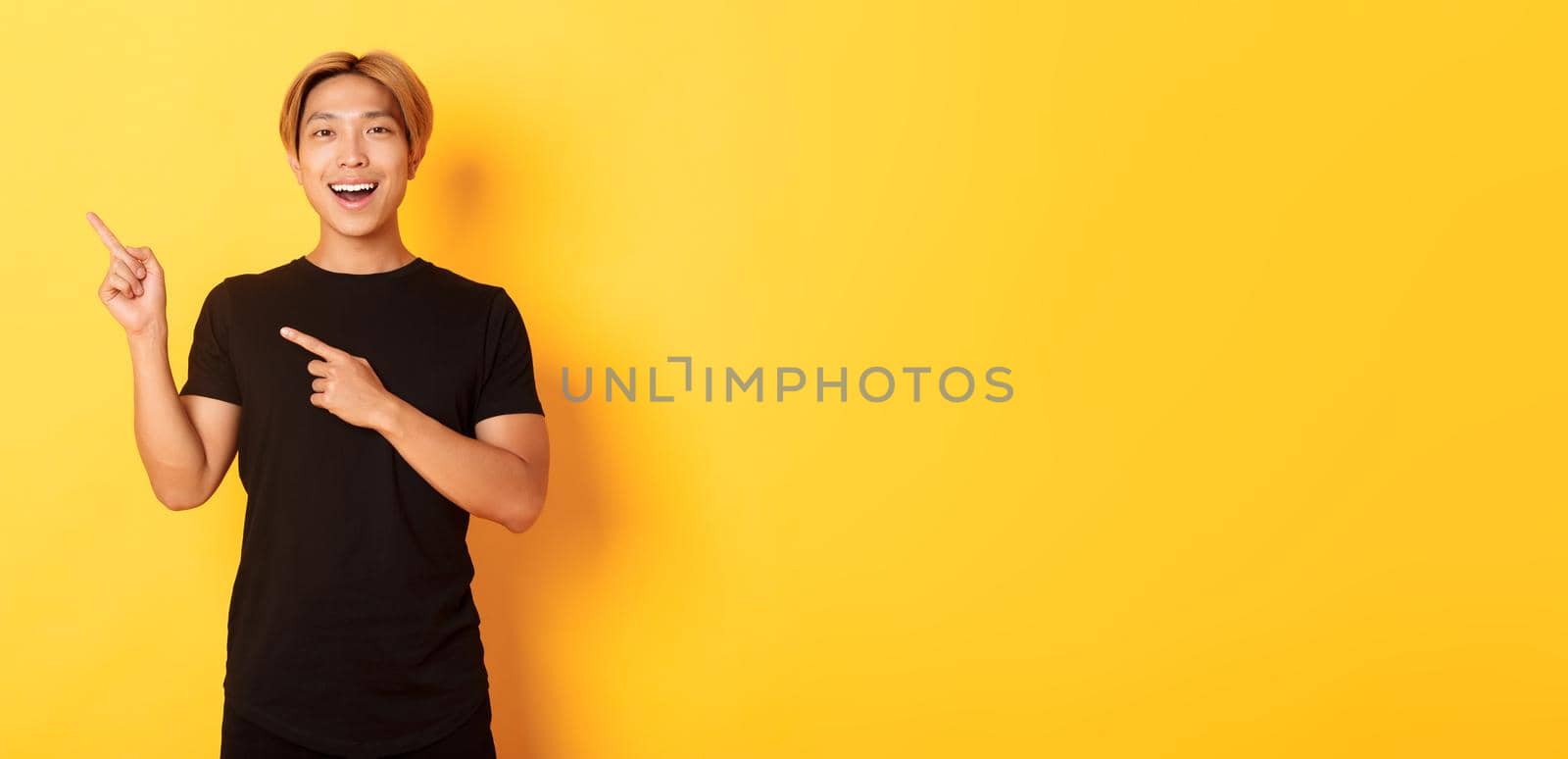 Portrait of cheerful asian man pointing fingers upper left corner, smiling and showing banner satisfied, yellow background by Benzoix