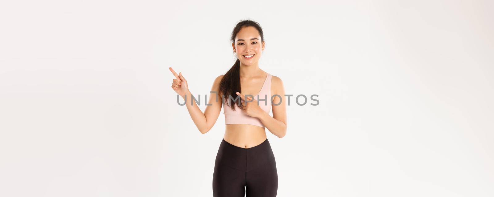 Sport, wellbeing and active lifestyle concept. Smiling fit and slim female fitness instructor, asian girl workout in gym and showing something, pointing fingers right, standing white background.