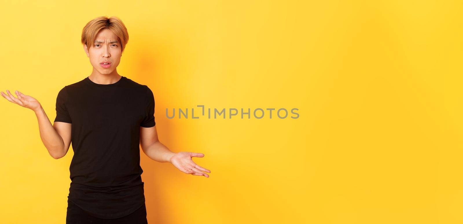 Portrait of confused and annoyed asian guy spread hands sideways, cant understand something, standing yellow background by Benzoix