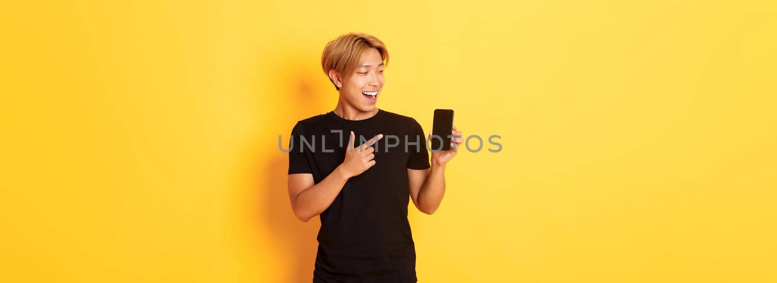 Portrait of satisfied asian guy pointing finger and looking at smartphone screen with pleased smile, showing application, standing yellow background.