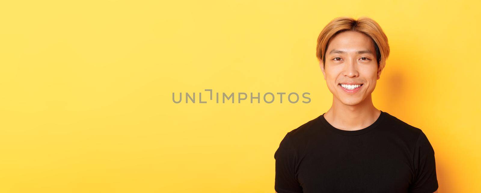 Close-up of handsome blond asian guy in black t-shirt, smiling happy at camera, standing over yellow background.