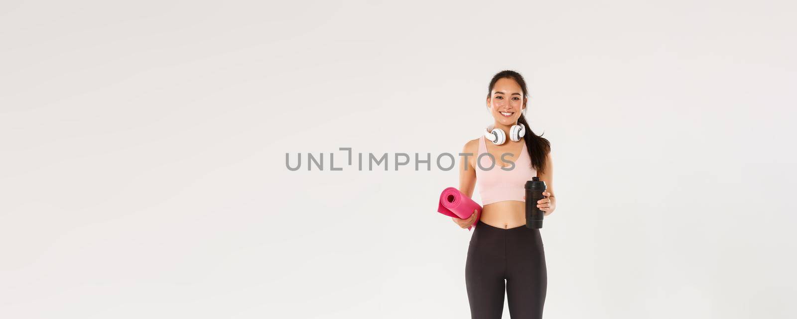 Full length of beautiful asian girl going for yoga classes, female athlete with headphones and water bottle smiling as carry fitness exercises rubber mat for her training in gym, workout concept.