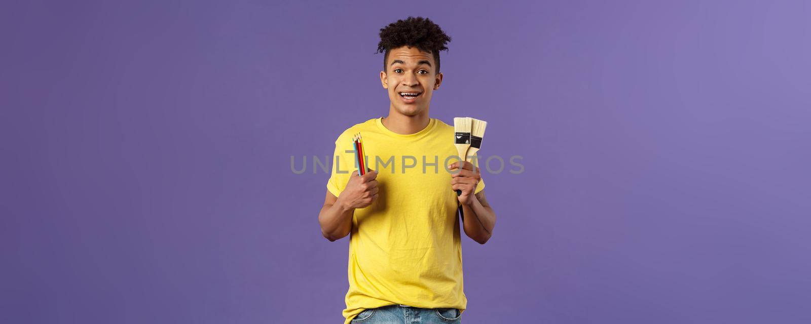 Portrait of upbeat young creative man, artist starting art courses, want to learn how to draw, holding colored pencils and brushes, standing enthusiastic over purple background.