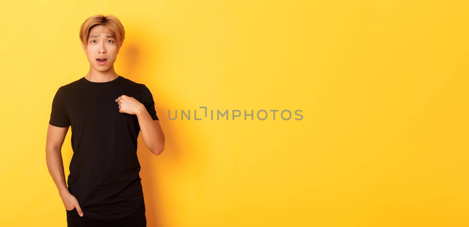 Portrait of confused and sad asian blond guy, frowning upset and pointing at himself, standing yellow background.