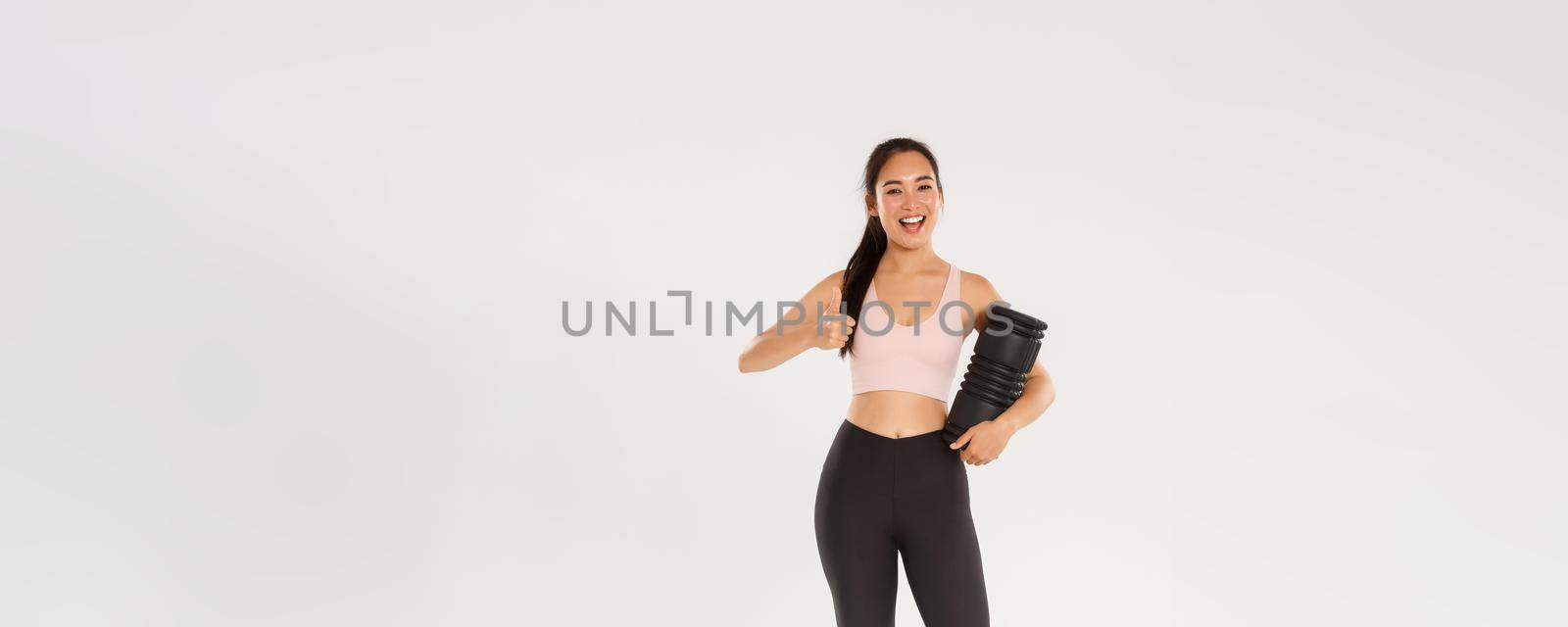 Full length of satisfied smiling asian female training coach, fitness girl in sportswear looking pleased, showing thumbs-up, using foam roller after workout to reduce soreness and ache in muscles by Benzoix