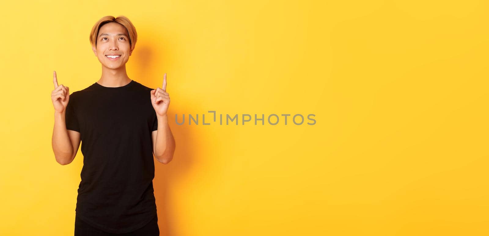 Handsome smiling asian man in black t-shirt pointing fingers up, looking at banner pleased, yellow background.