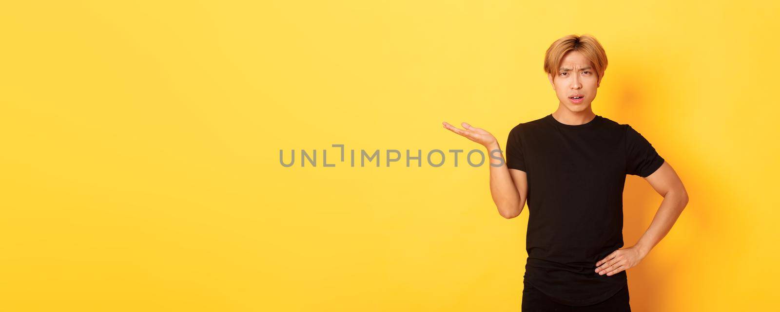 Frustrated and annoyed asian guy with blond hair, raising hand confused and looking at camera, yellow background.