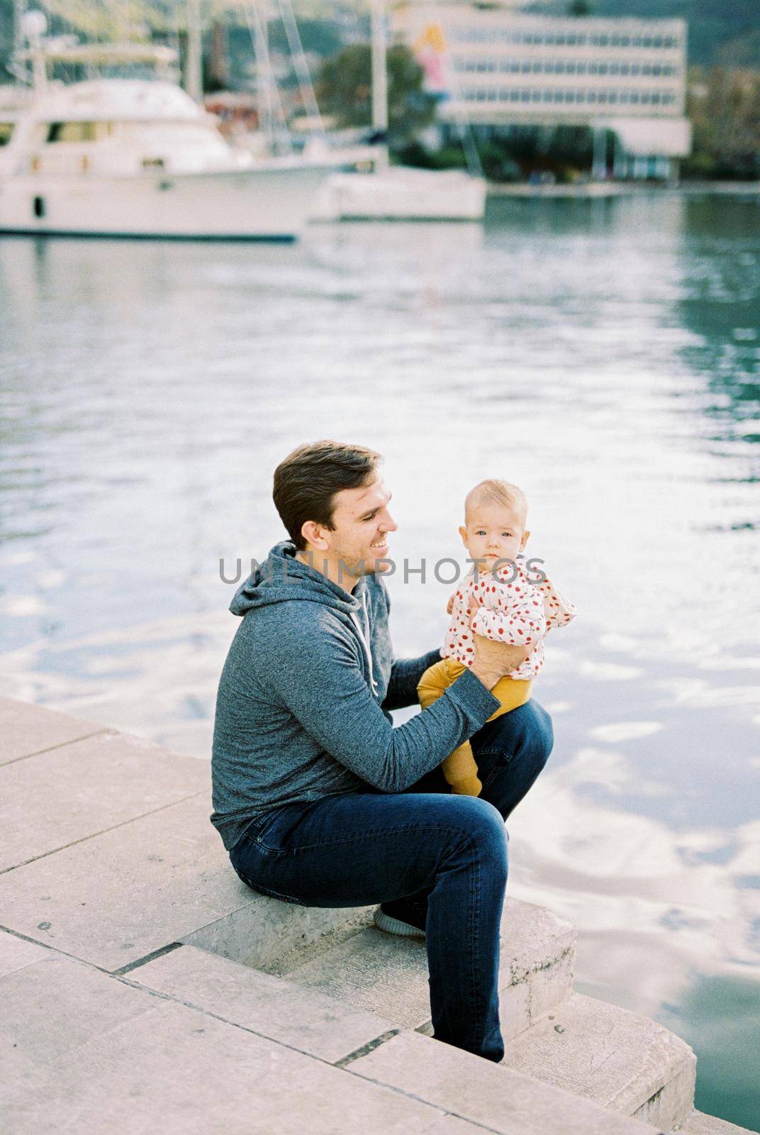 Dad sits on the pier with a baby on his lap against the backdrop of yachts by Nadtochiy