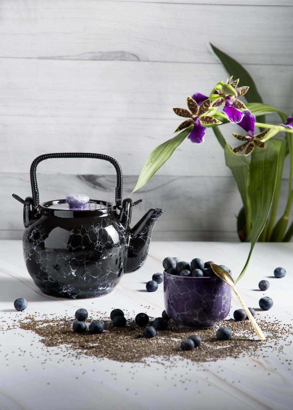 still life with a black ceramic teapot and a cup of tea with blueberries on a light background with an orchid, chia seeds and berries are scattered on the table,High quality photo