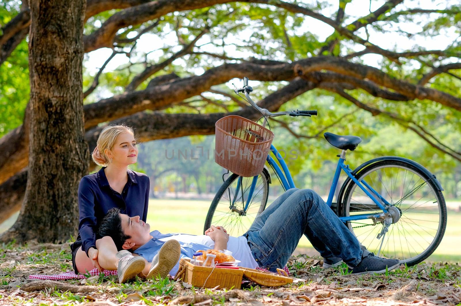 Beautiful girl sit with her boyfriend who recline beside and bicycle is on the back under the big tree in the garden.