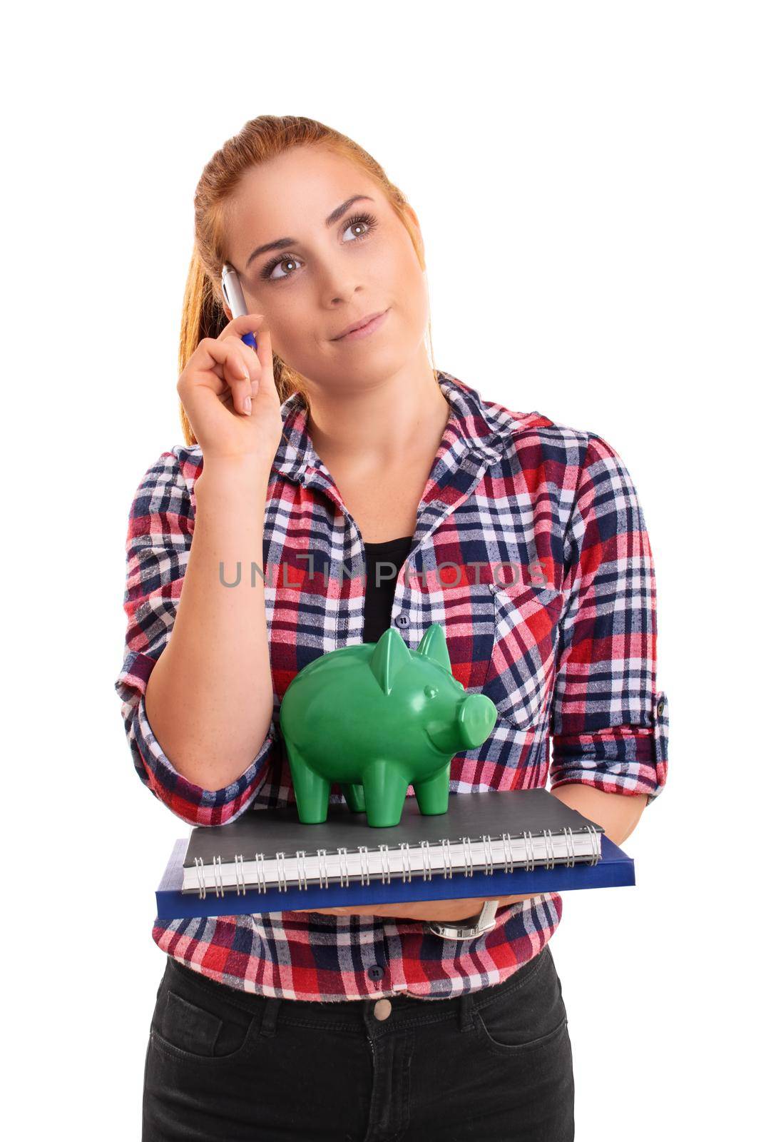 Young female student holding a green piggy bank on a stack of books, thinking with a pen on forehead, isolated on white background. Finance, banking, loan, savings concept.