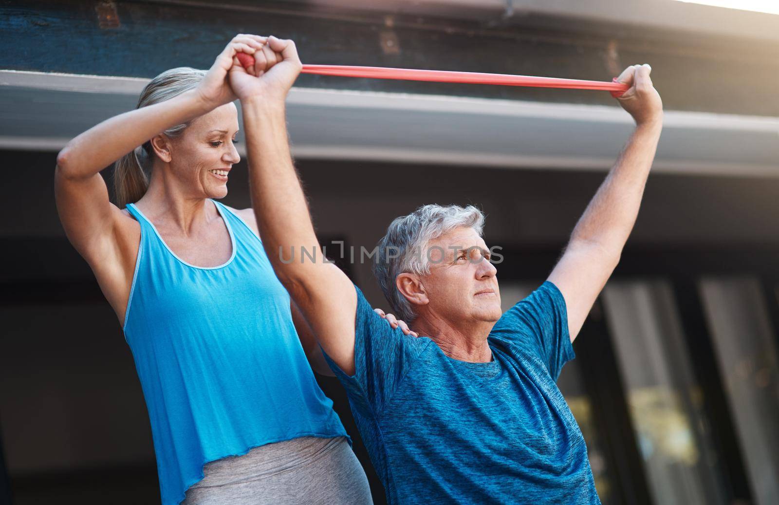 She always gives me that extra push. a mature man stretching his arm muscles with an elastic band while being assisted by his wife. by YuriArcurs