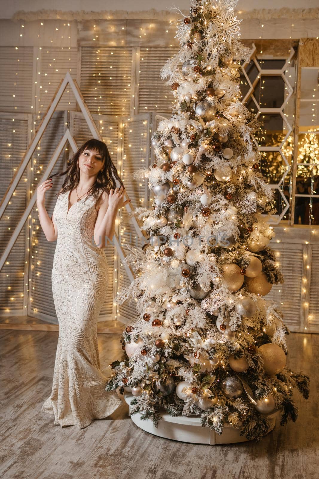 A brunette woman in a beautiful light tight dress next to a Christmas tree. The magical atmosphere of Christmas Eve on December 25. Decoration for the holiday in a light style with lots of lights and light bulbs.