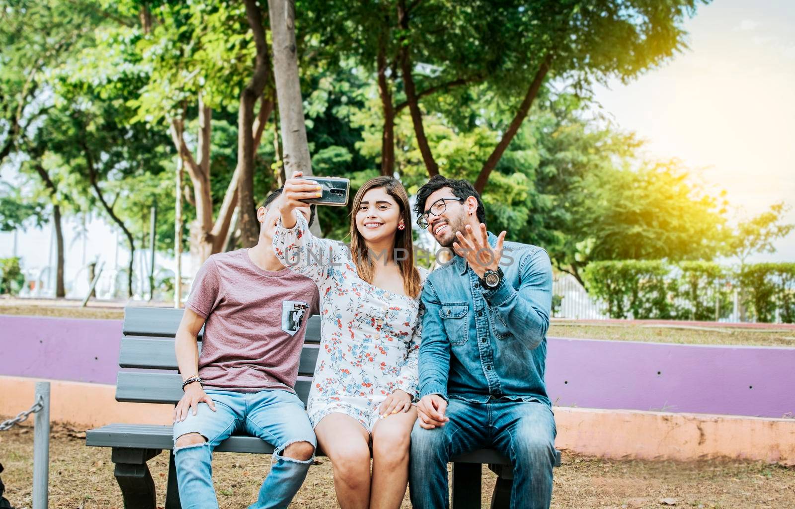 Three friends sitting on a bench taking a selfie, Three teenage friends sitting on a bench taking selfies. Gathering of three happy friends taking a selfie sitting on a bench by isaiphoto