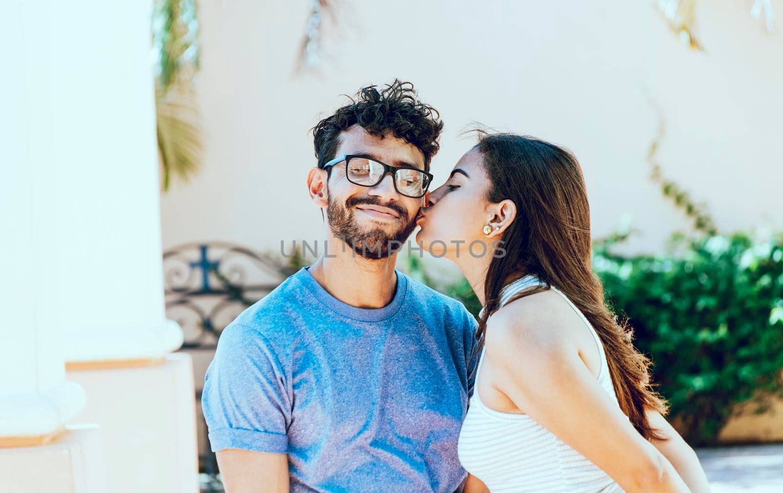 Girl kissing her boyfriend on the cheek while smiling. Young woman kissing her boyfriend on the cheek in the street. Concept of happy girlfriend kissing cheek of her boyfriend outdoors by isaiphoto