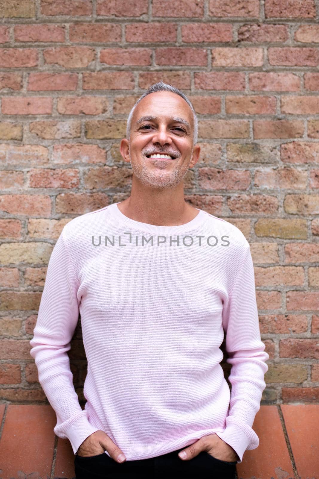 Portrait of smiling caucasian man wearing pink sweater looking at camera leaning against brick wall. Vertical image. Lifestyle concept.