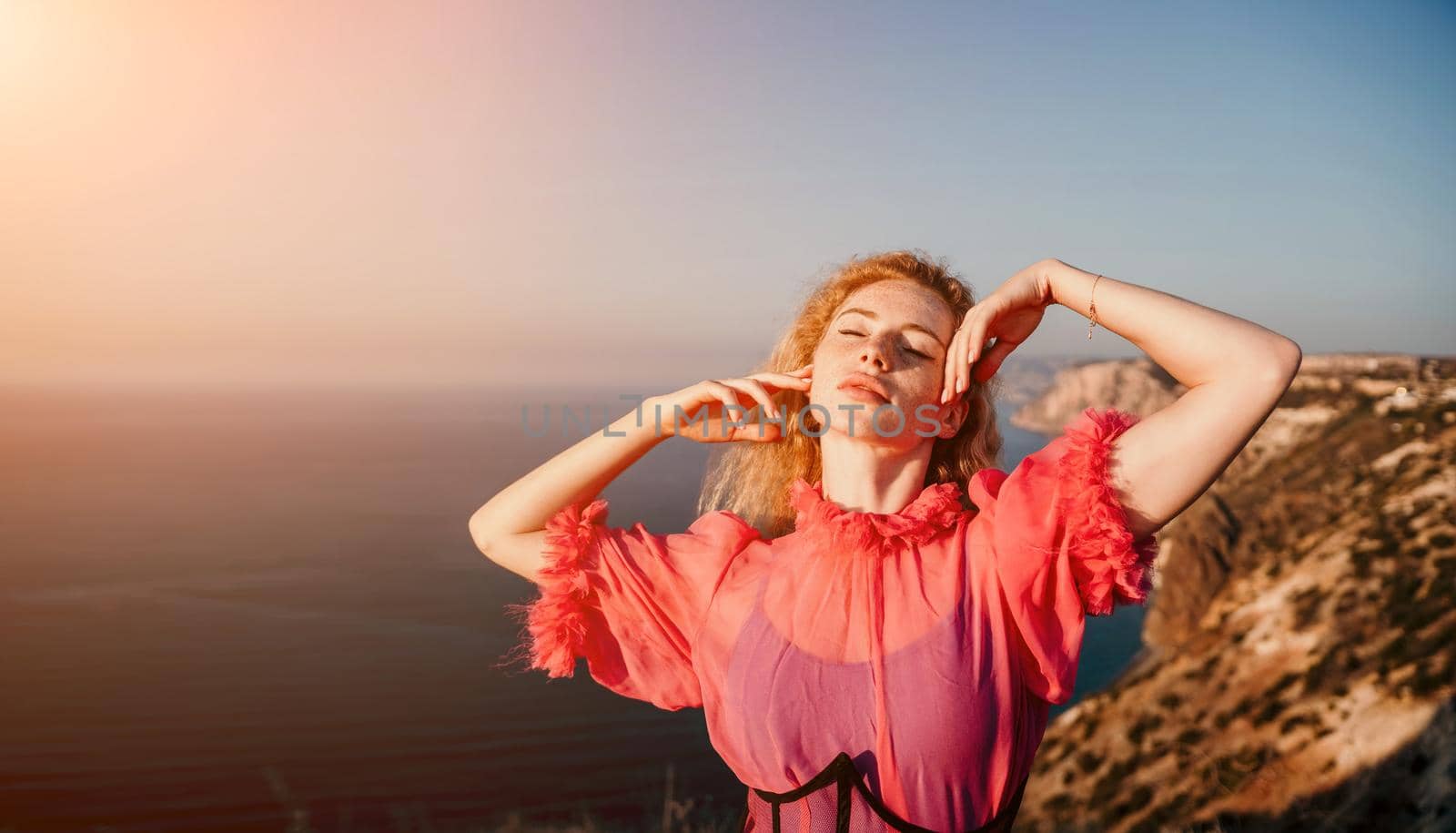 Close up shot of beautiful young caucasian woman with curly blond hair and freckles looking at camera and smiling. Cute woman portrait in a pink long dress posing on a volcanic rock high above the sea